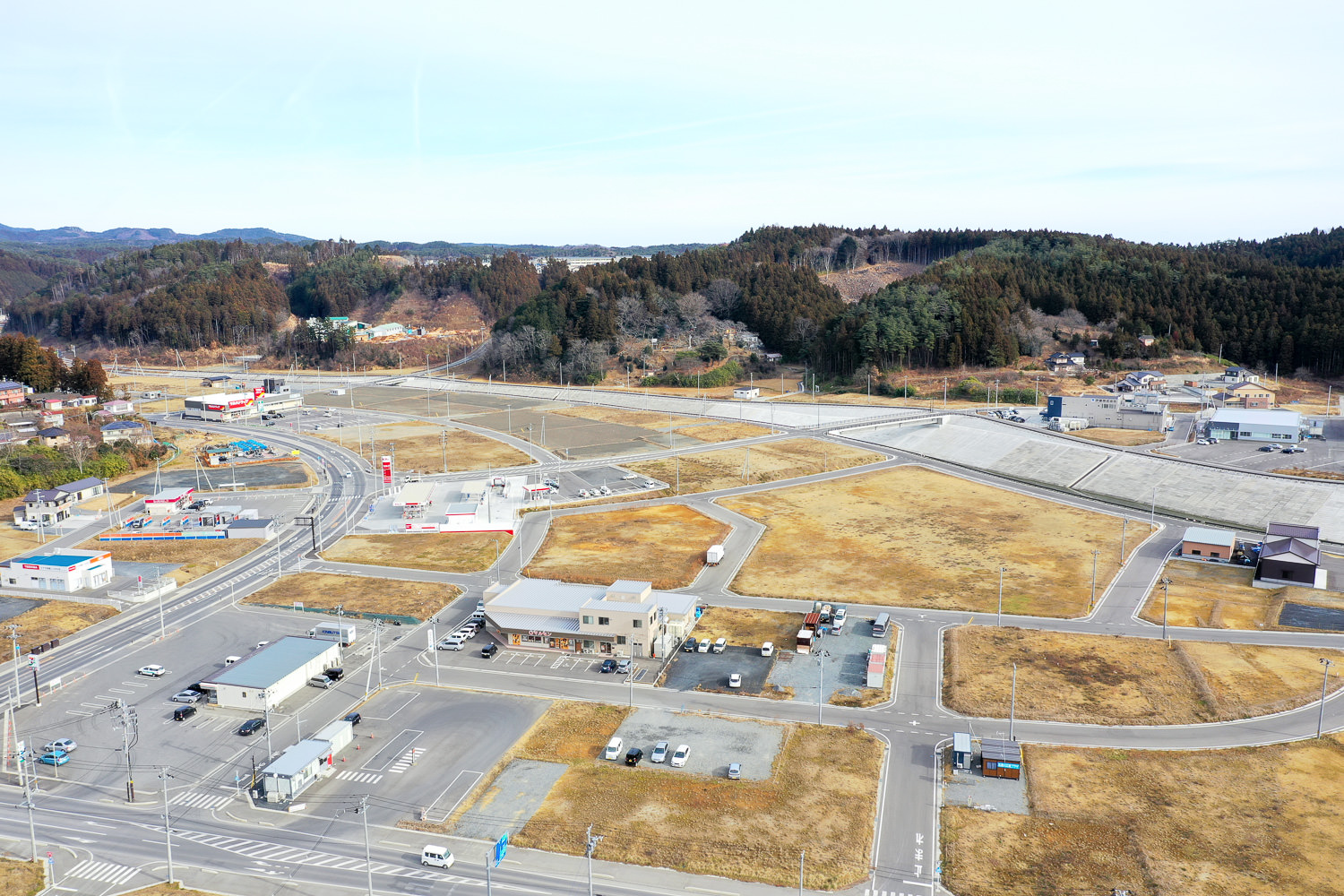晴れた空の下、建物や道路、駐車場が点在する準都市的な地域を上空から捉えた風景で、草地や空き地が広がり、背景には森林に覆われた丘が見える写真
