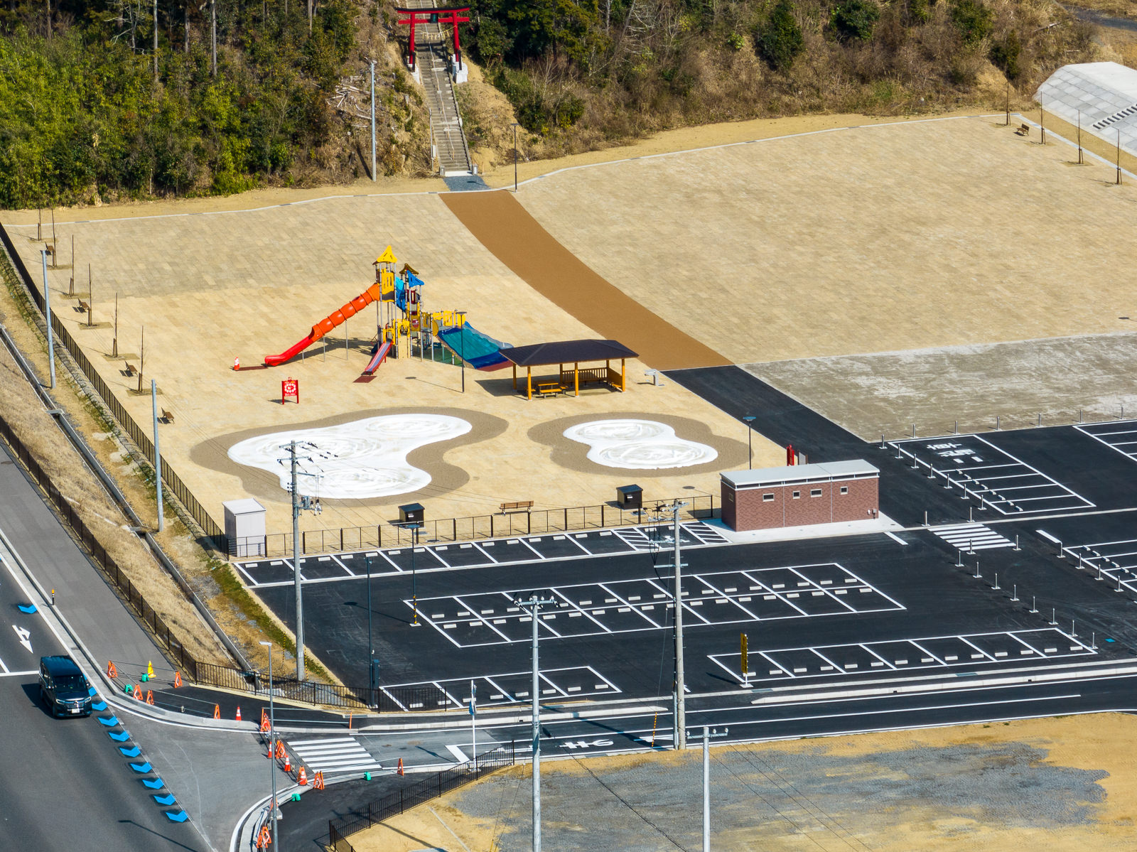 広い駐車場に隣接して明るい色の地面に複合遊具や東屋などが設置された新しい公園があり、奥の斜面には神社へと続く赤い鳥居と長い石段が見える、山のふもとに整備された子ども向け遊び場の昼間の風景を高台から定点カメラで撮影した写真