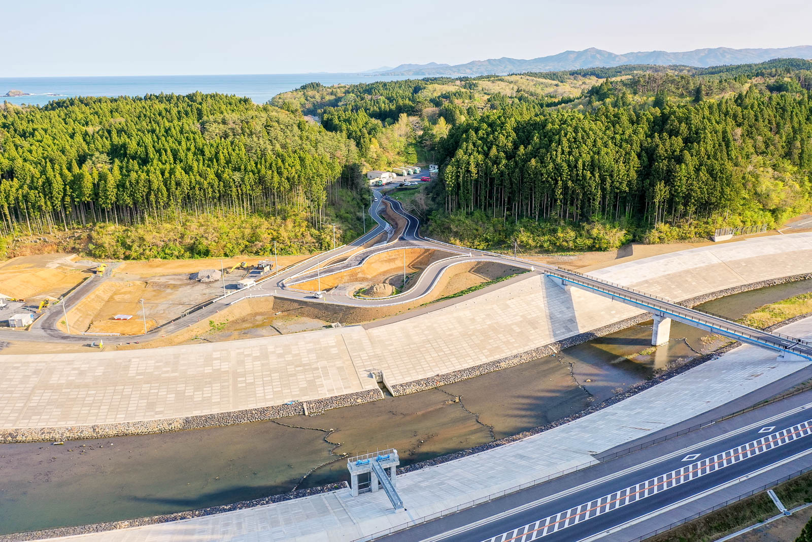 手前にコンクリートで固められた護岸とその間を流れる川、中央に川をまたぐ橋と整備中の道路、その奥には密集した杉林と山並みが広がり、背景に海と遠くの陸地が見える、自然と人工構造物が共存する復興途中の沿岸地域を晴天下で撮影した写真