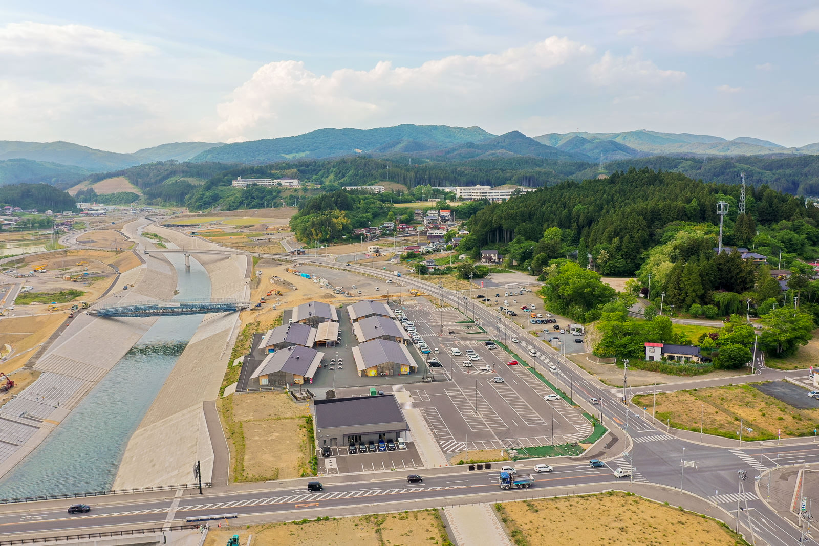 川が町の中央を流れ、周囲に建物、駐車場、道路が広がり、背景には緑の丘と山々が見える小さな町の空撮写真