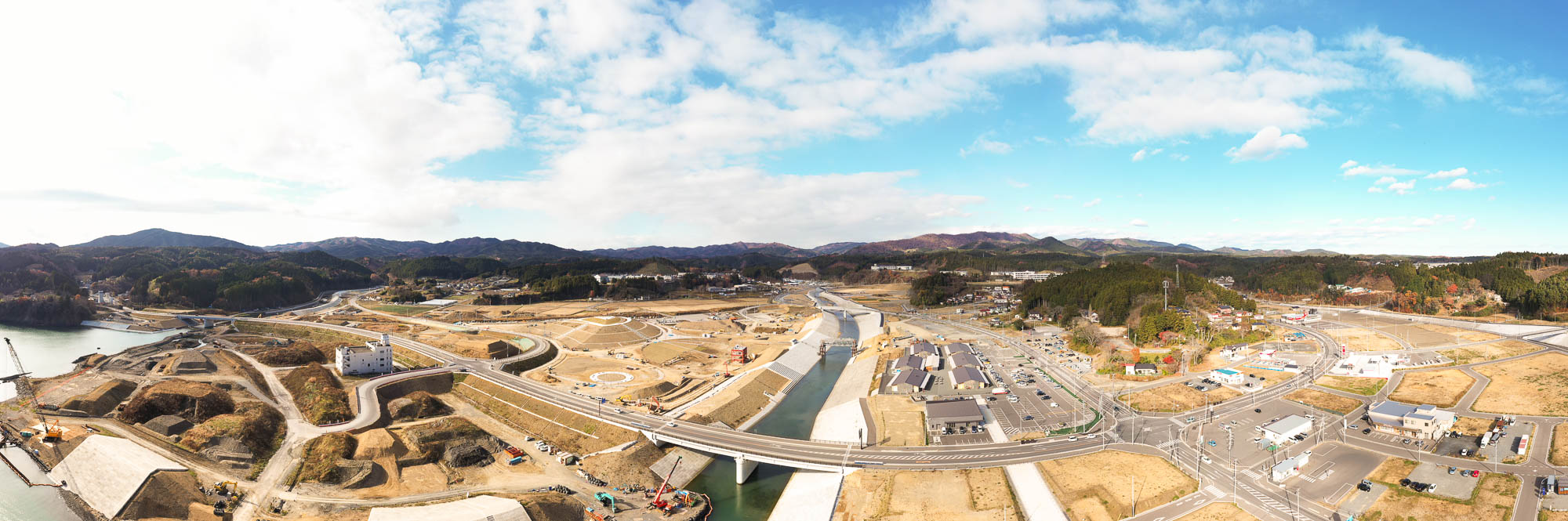 山に囲まれた広い造成地の中央を川が流れ、複数の道路と橋が交差し、周囲に工事車両や建物が点在している様子を上空から広く見渡した写真