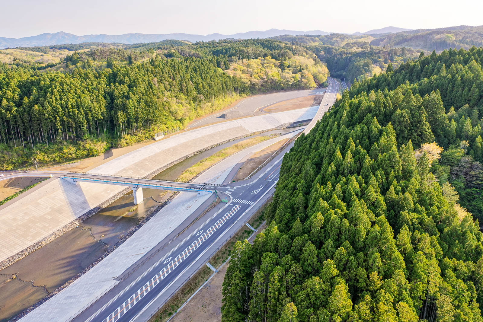手前に青々とした杉の森林が広がり、中央には川にかかる橋と護岸が整備された河川沿いの風景、その横を走る新しい道路、奥にはなだらかな山々と自然豊かな景色が広がる様子を高所から撮影した、山間部の復興インフラが整備された地域の写真