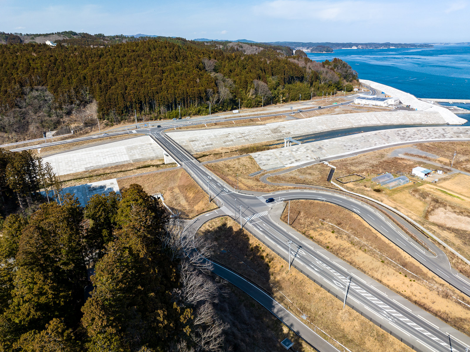 山林を背にして、防潮堤に沿って新しく建設された道路と橋が交差し、周囲には農地と小さな建物が点在している、海辺に近いインフラ整備の昼間の風景を高台から定点カメラで撮影した写真