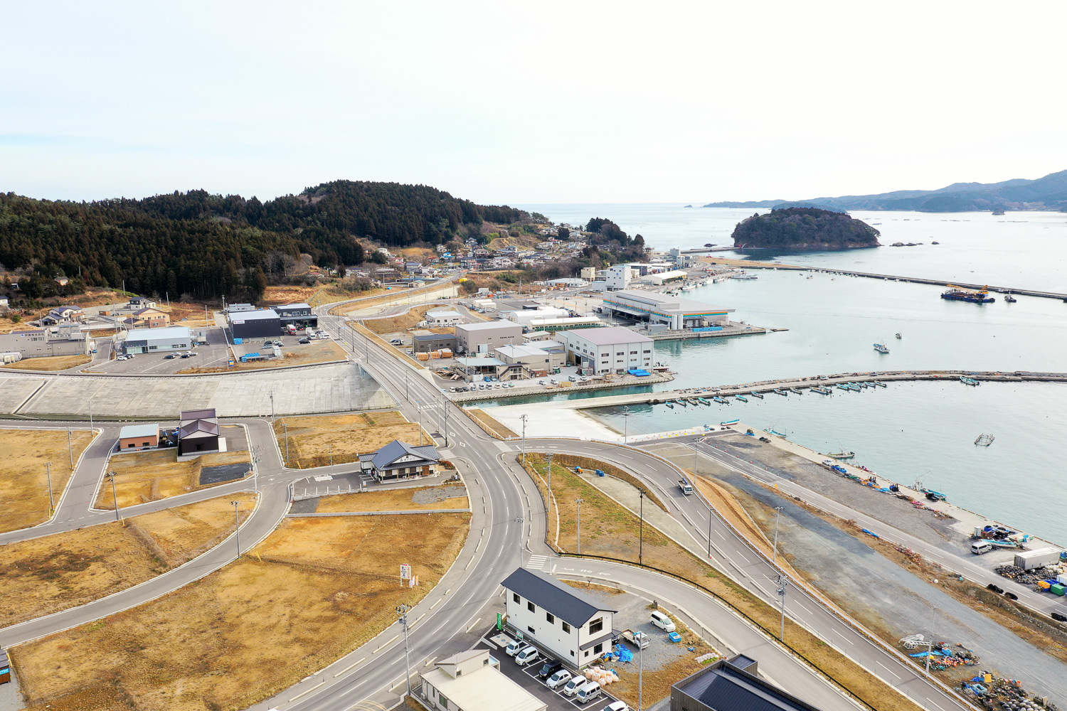 海沿いの町を上空から撮影した写真で、複数の道路が交差しながら建物や家屋が点在し、港には船が停泊している様子が広がっている海沿いの町の写真