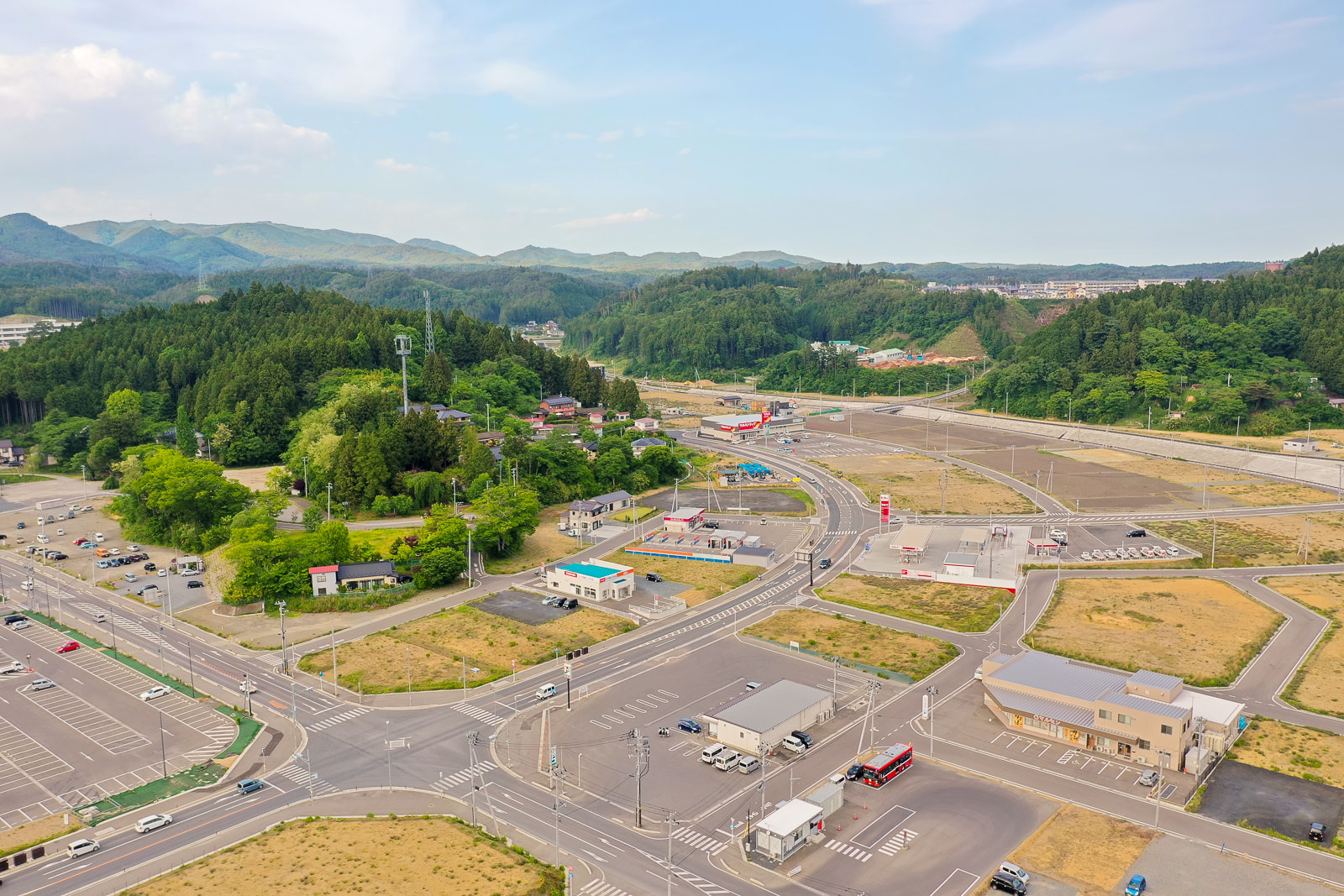 広い駐車場と交差点があり、周囲には緑豊かな山々と建物が点在している風景の写真