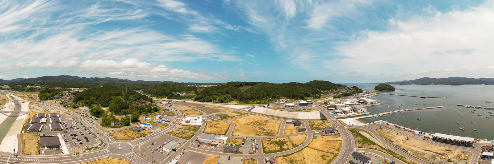 海に面した町を広く見渡し、建物や道路、緑地が広がり、海には複数の船が浮かび、背景には丘と森林が広がる、雲が少し浮かぶ空の下の沿岸の町の写真