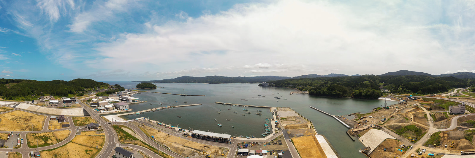 港に複数の船が浮かび、海岸沿いに建物やインフラが並び、自然の緑と開発地域が混在し、背景には丘が広がる、雲が少し浮かぶ空の下の沿岸地域の写真