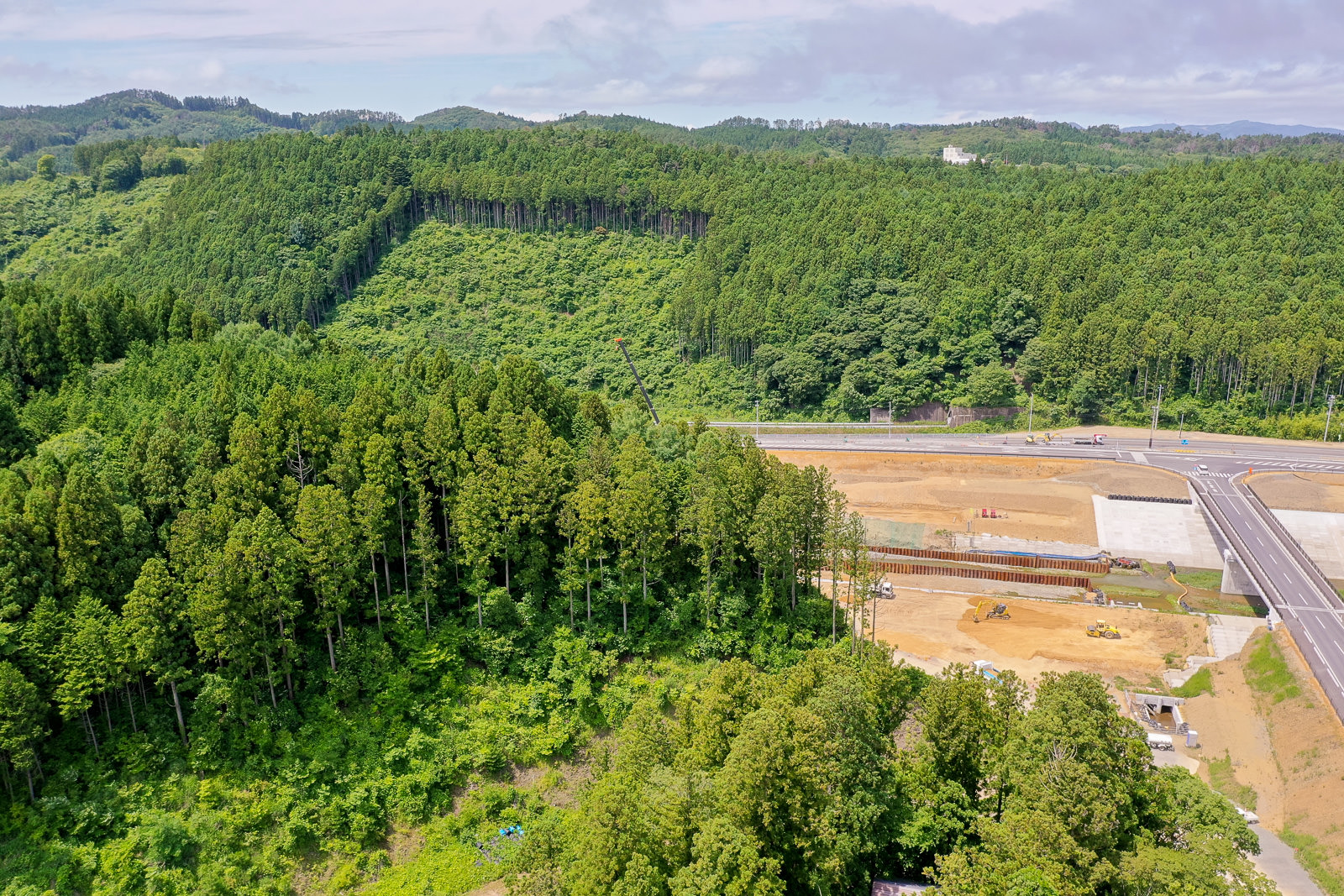 背景に緑豊かな丘陵地帯が広がり、手前には重機や建設中の構造物がある造成地と、それに沿って走る道路が見え、空には雲が少し浮かぶ風景の写真