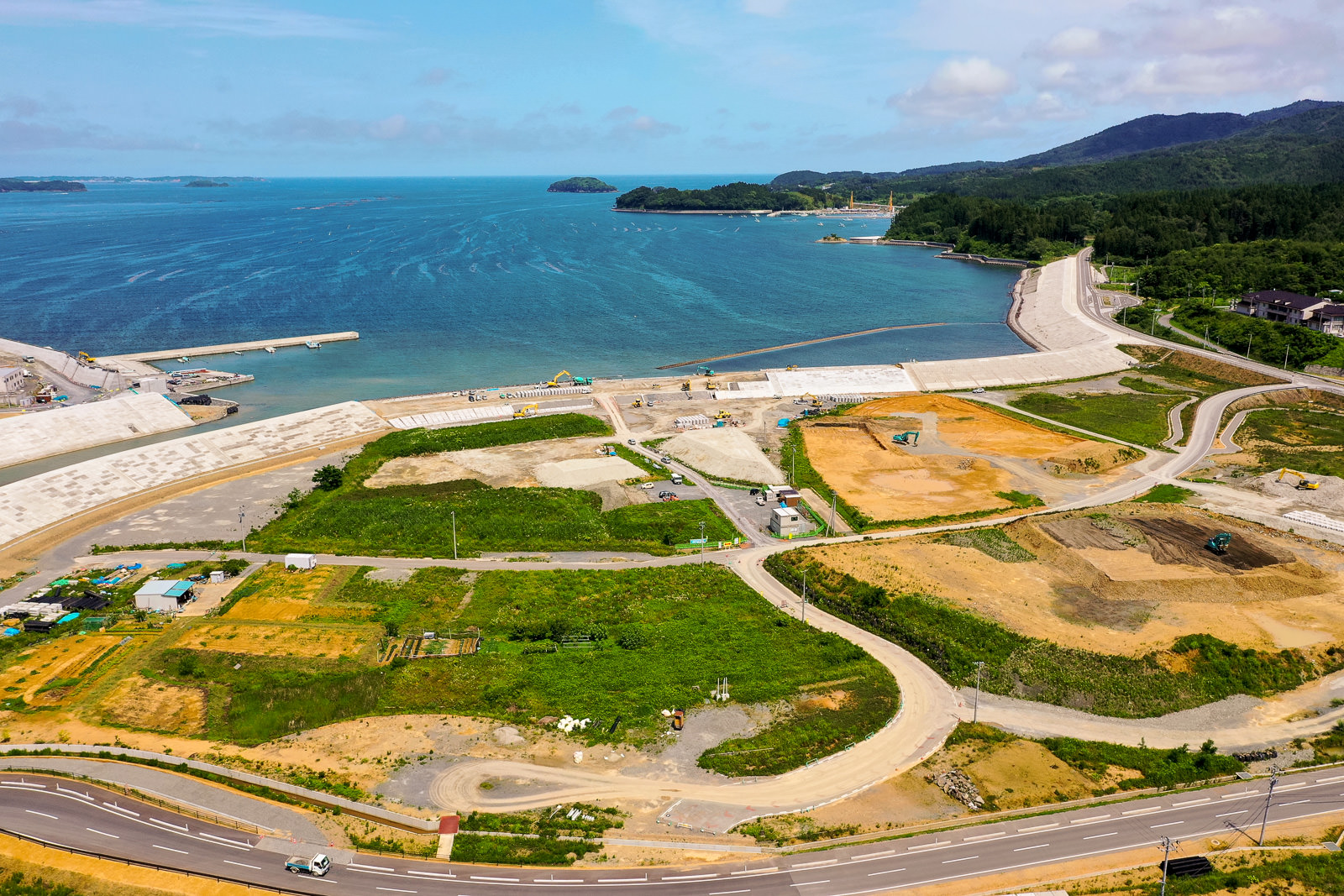緑の草地と工事現場、道路が広がる沿岸地域を空から見下ろし、背景には海と小さな島々が見え、海岸線にはコンクリート構造物が設置され、複数の建設車両や機材が点在する晴れた空の下の写真