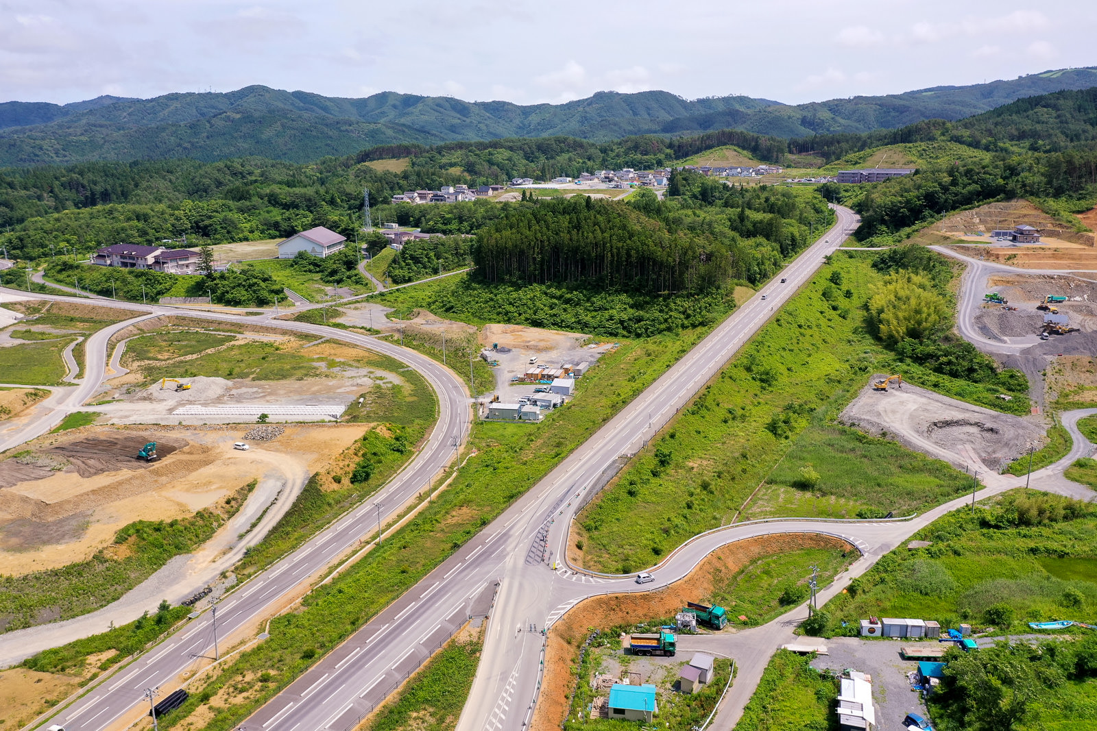 緑の丘と森林が広がる田舎の風景の中で、複数の舗装された道路が交差し、一部に車両が走行しており、背景には建物が点在する集落と山々が見える晴れ時々曇りの空の下の写真