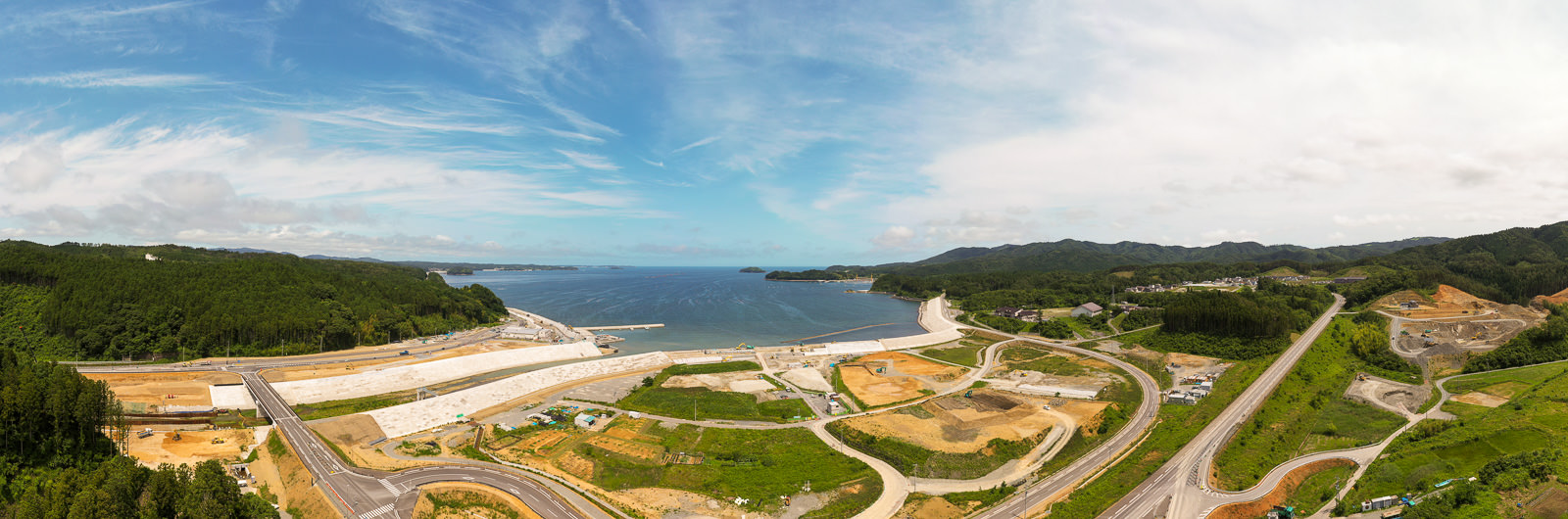 中央に広がる海を背景に、道路や歩道、工事現場のような開発区域が点在し、両側には緑の丘と森林が広がり、空には雲が少し浮かぶ沿岸地域の写真