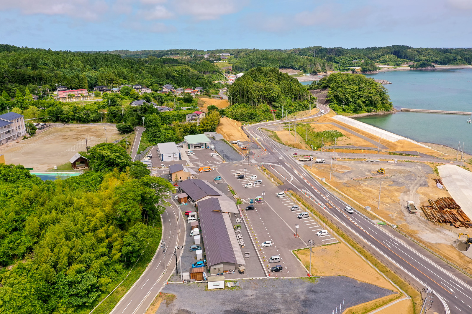広い駐車場に複数の車が停まっており、長い屋根の建物や道路が周囲に広がり、右側には海岸線とその近くで進行中の開発工事が見られ、背景には緑豊かな森林や丘、点在する住宅や構造物が広がる空撮の写真