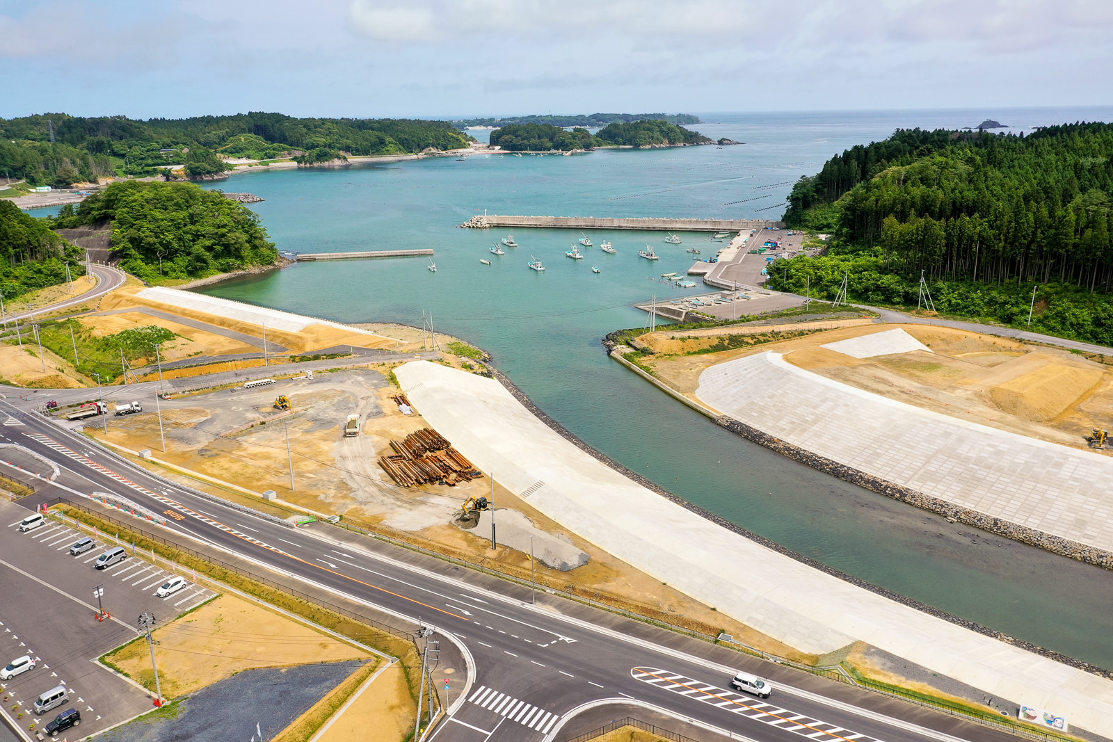 海沿いの港に複数の船が停泊しており、手前には道路や駐車場が広がっていて車両が停まっており、広く整地された場所や建設資材が見られる開発中の様子で、背景には緑豊かな丘と木々、そして遠くに広がる海が見える写真