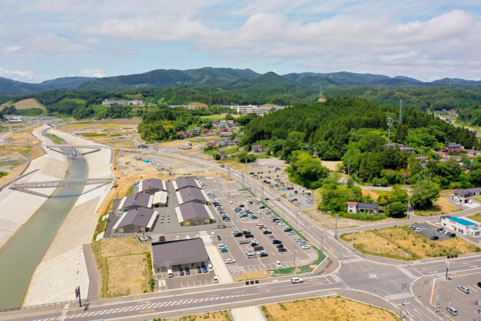 川が中央を流れ、両岸にコンクリートの護岸が施され、橋が架かる町を空から見下ろし、建物や駐車場、車が並び、背景には緑の丘と山々が広がる写真
