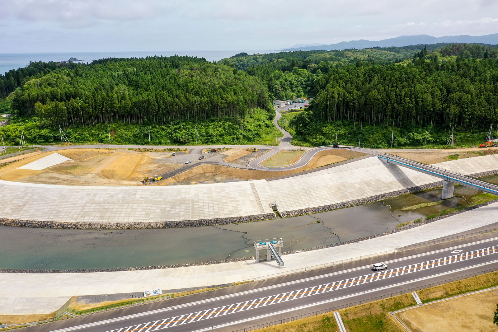 川の両岸にコンクリートの護岸が整備された風景を上空から撮影しており、川には橋がかかり、手前には川に沿って道路が走り、背景には森林に覆われた丘と一部開発中の土地が広がる写真