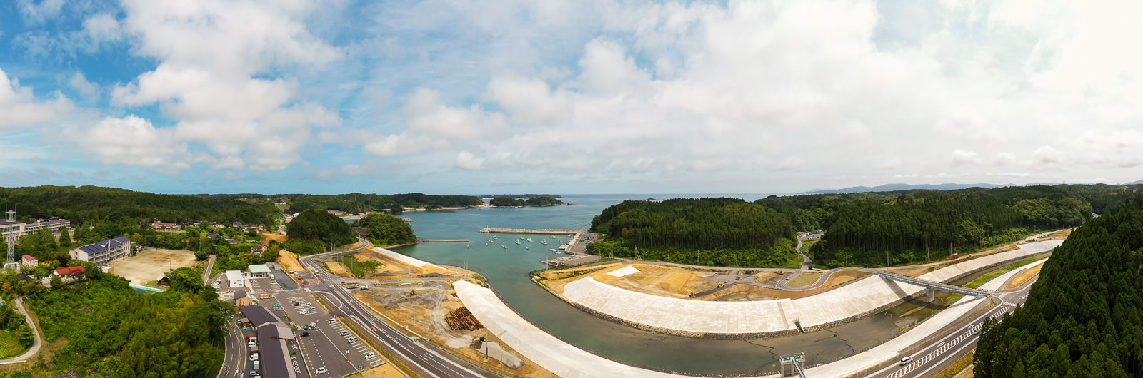 中央に入り江のような水域が広がり、その周囲を緑豊かな森林と一部整地された土地が囲み、道路やインフラが整備されていて、水辺には堤防のような構造物が見られ、空には青空と雲が混在する風景の写真