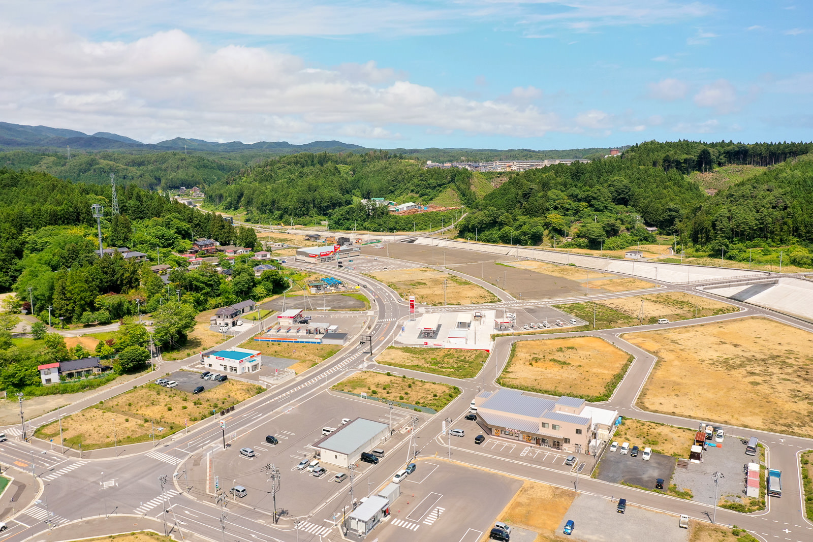 建物や道路、駐車場が点在する半都市型の地域を空から見下ろし、周囲には緑地が広がり、背景には丘が見え、空には雲が少し浮かぶ風景の写真