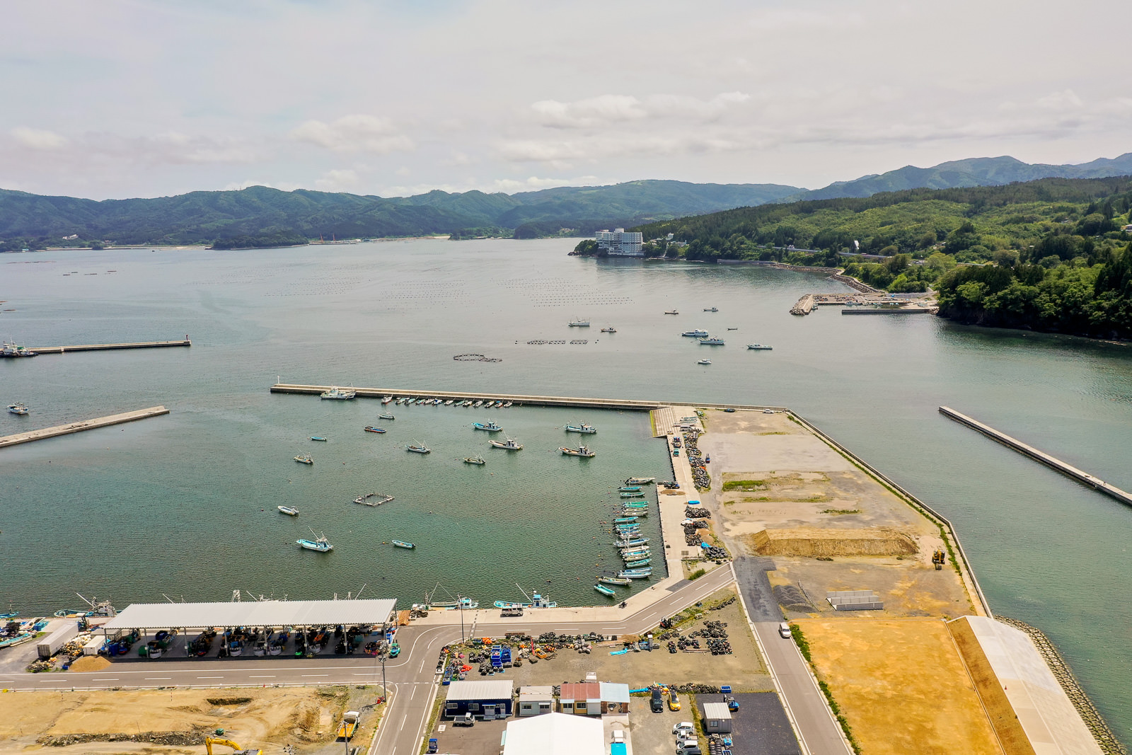 海に面した港で、複数の船が停泊しており、岸壁にはクレーンや倉庫のような建物が並び、遠くには山が見える風景の写真