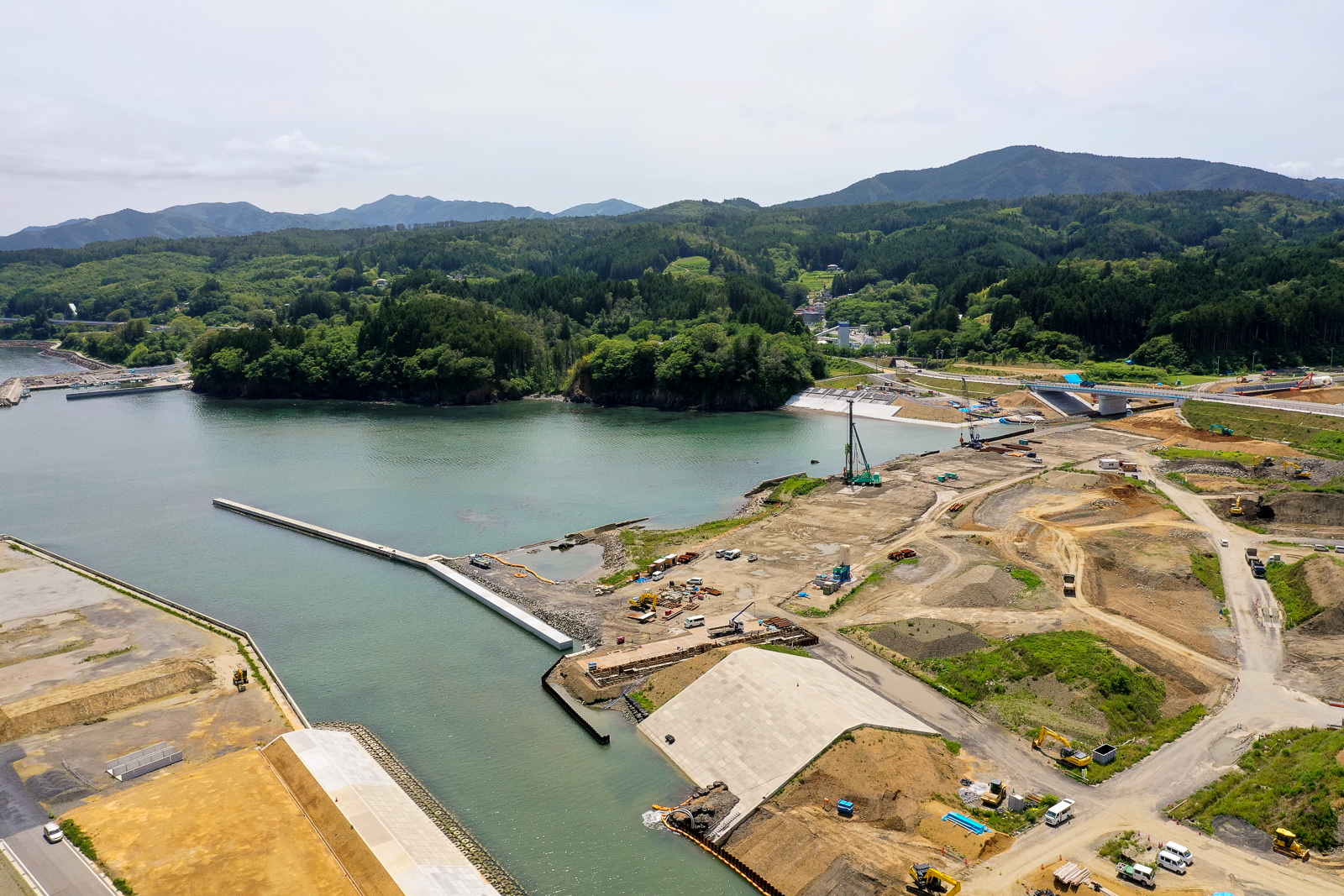 複数のクレーンやトラックなどの建設車両が稼働している海沿いの工事現場で、広い土地が整地されて建設準備が進められており、コンクリート構造物が海に向かって延びていて、背景には緑豊かな丘や森林が広がる写真