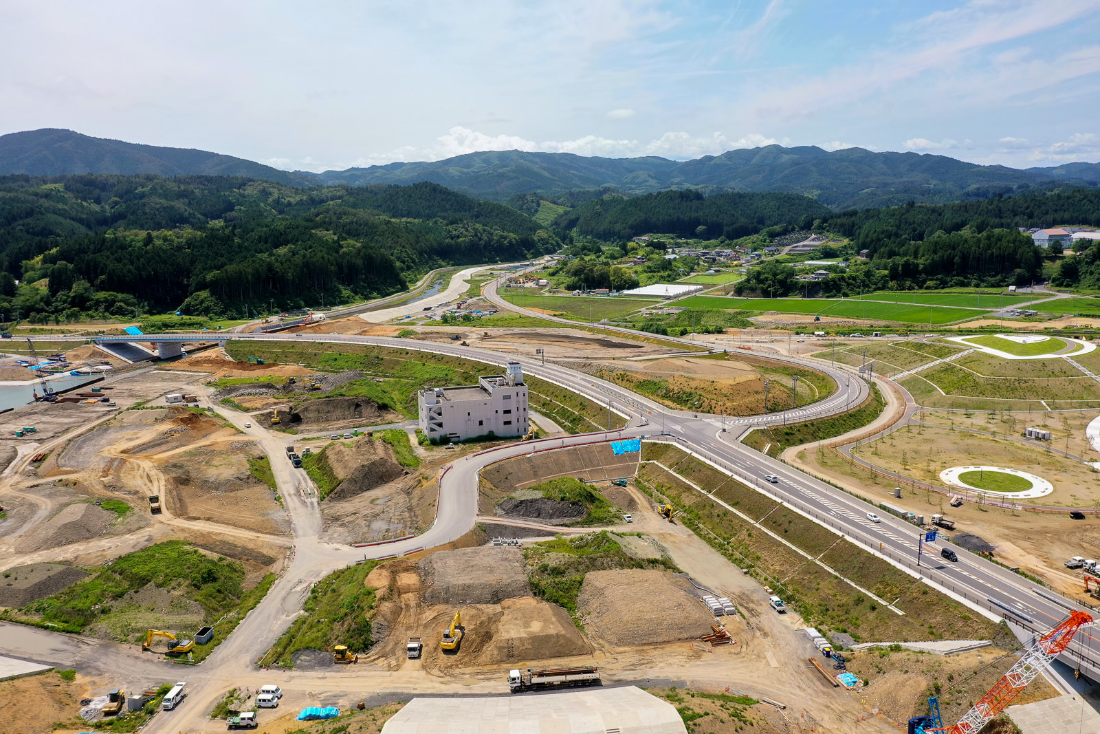 山に囲まれた広大な土地で、道路や建物が建設中の様子を上空から撮影しており、重機や資材が点在し、開発が進む様子が一望できる写真