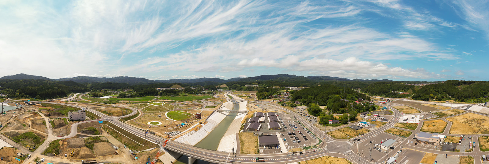 中央を川が流れ、両岸にコンクリートの護岸が施され、その周囲に道路や建物、駐車場が広がり、所々に工事現場や建設機材が見られ、背景には山々がそびえ、空には薄い雲が浮かぶ開発中の地域を俯瞰した写真
