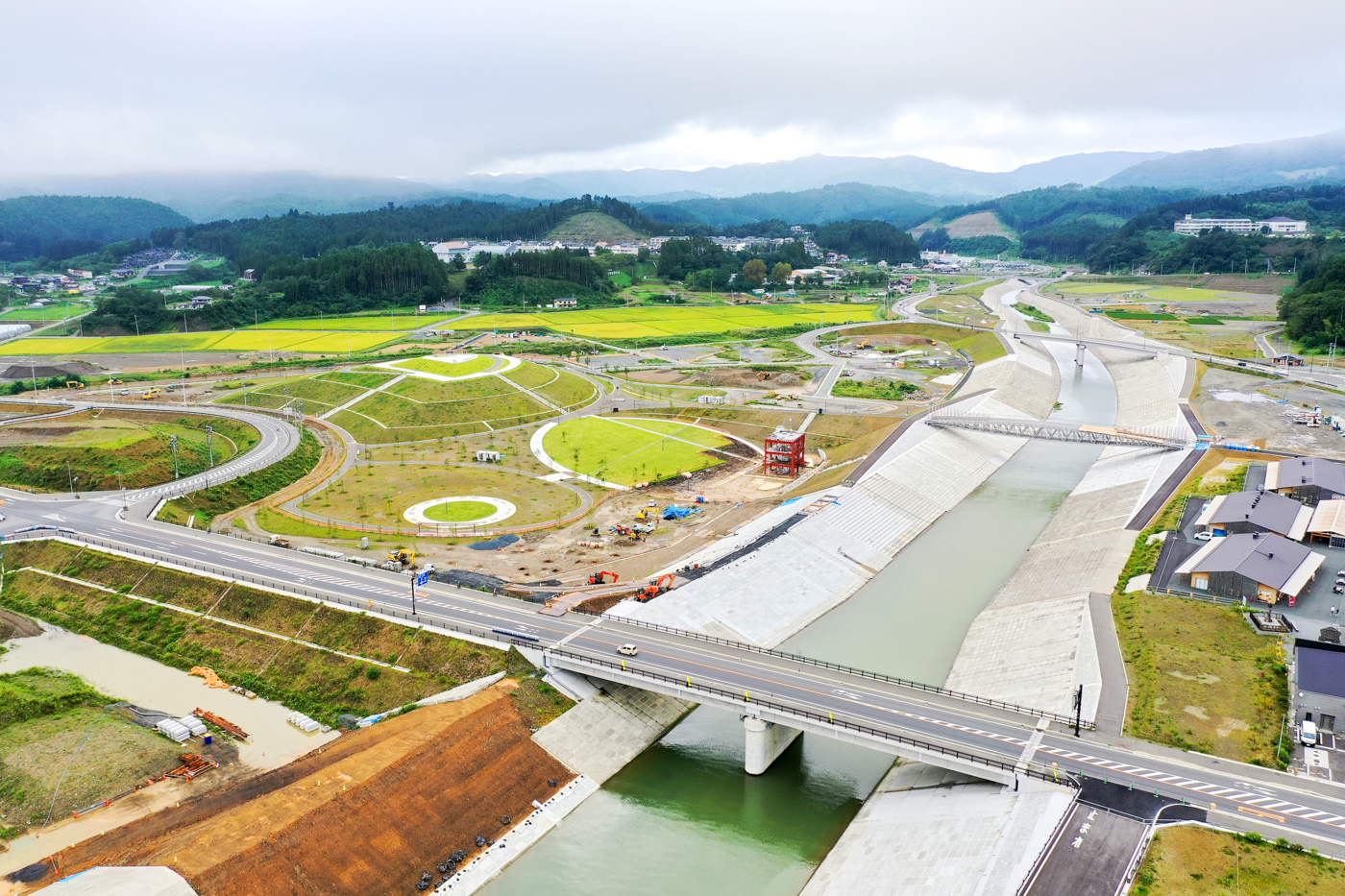 広々とした田園風景の中をゆるやかに川が流れており、その川の上にはコンクリート製の橋が架かっていて、周囲には緑の畑や草地が広がっている穏やかな風景の写真