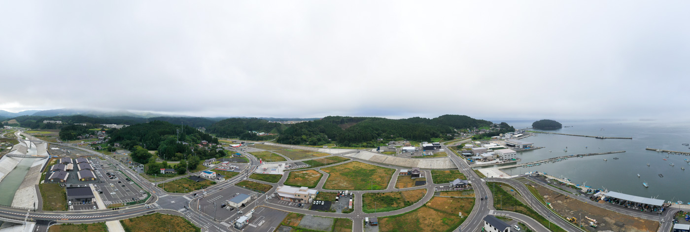 海沿いに広がる町を上空から撮影した風景で、複雑に交差する道路の周囲に建物や駐車場が点在し、右側には港が広がり複数の船が停泊している様子が見える海沿いの町の写真