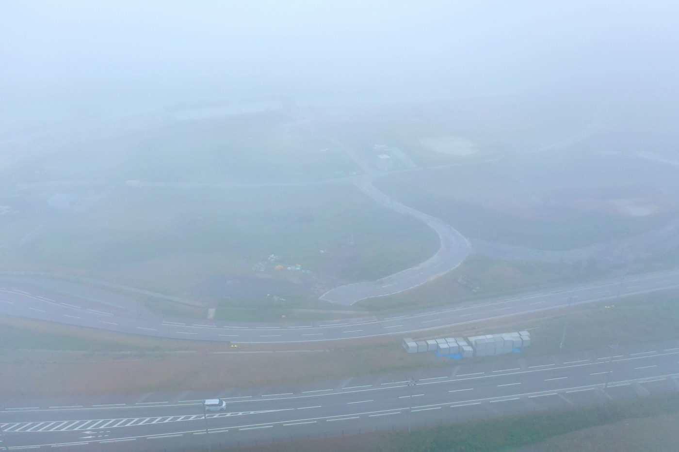 濃い霧に包まれた風景の中で、舗装された道路が静かに伸びており、周囲の木々や地形が白くかすんで見える幻想的な雰囲気の霧の写真