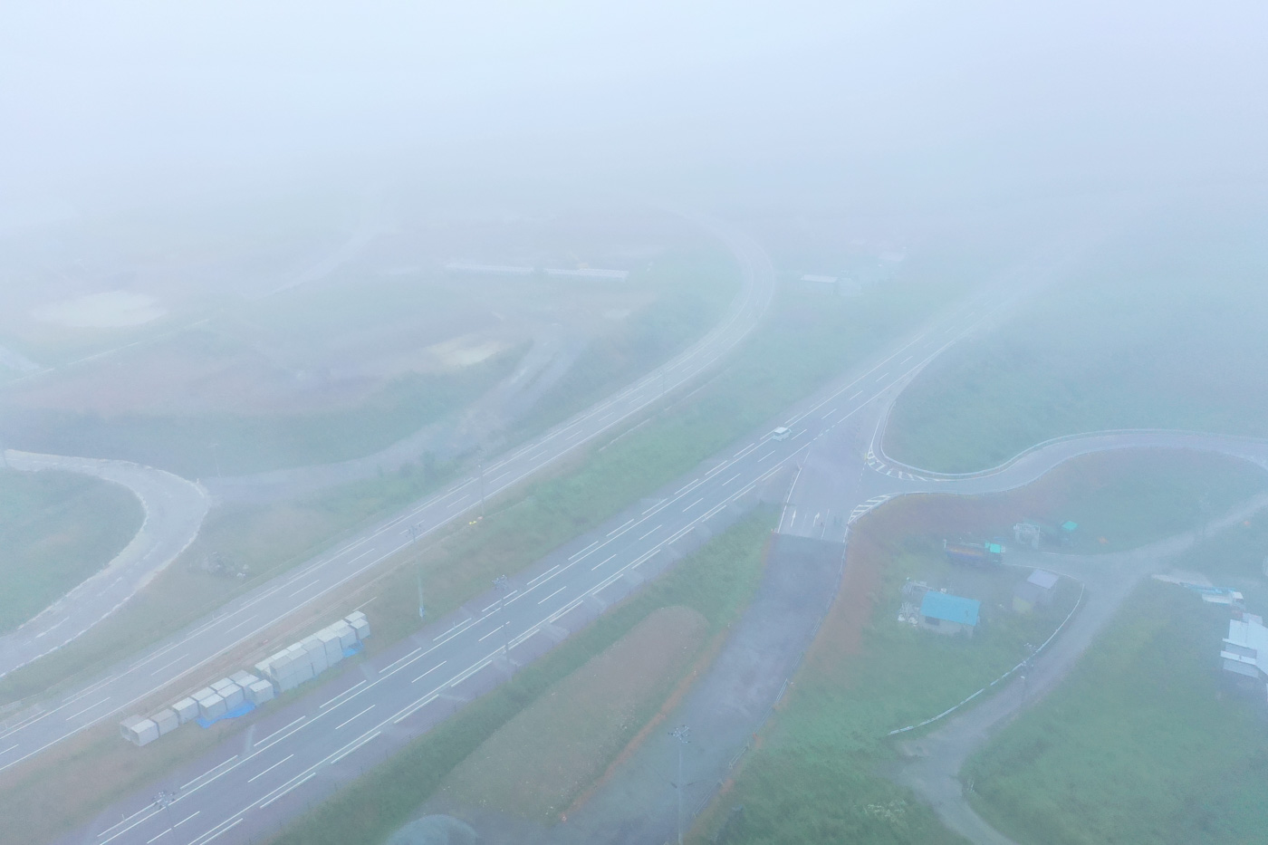 濃い霧に包まれた風景の中で、曲がりくねった道路が交差点に向かって伸びており、信号機や標識がかすかに見える様子が写された交差点の写真