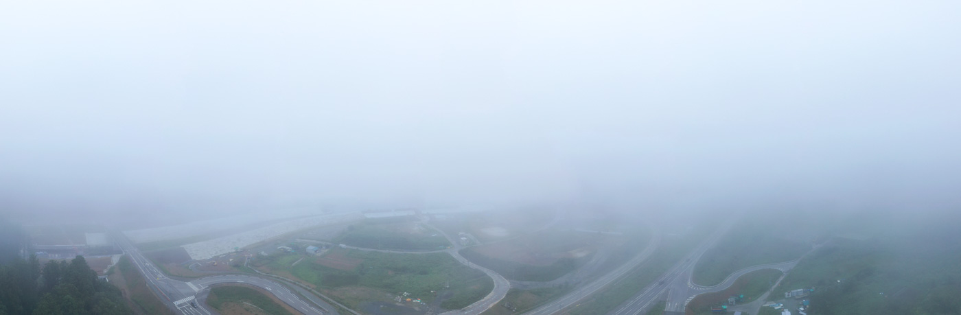 霧が立ち込める中、視界がぼんやりとした道路がまっすぐに伸びており、周囲の風景がかすんで見える静かな様子が写された道路の写真