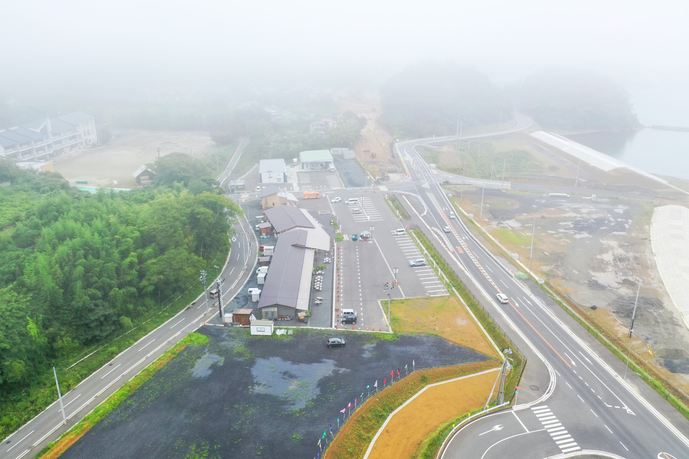 霧が立ち込める風景の中で、複数の道路に囲まれた広い駐車場とその隣に建物が並んでおり、周囲の景色が白くかすんで見える静かな町の一角の写真