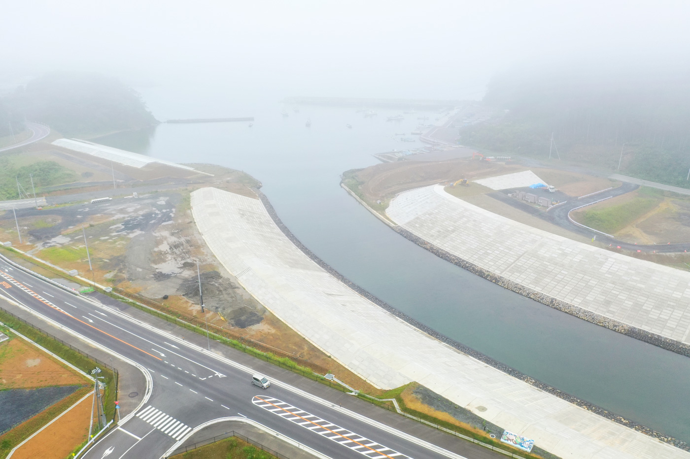 霧が立ち込める湾の周囲に護岸工事が施された地域が広がっており、その手前には複数の道路が交差する交差点が見える静かな海辺の工事現場の写真