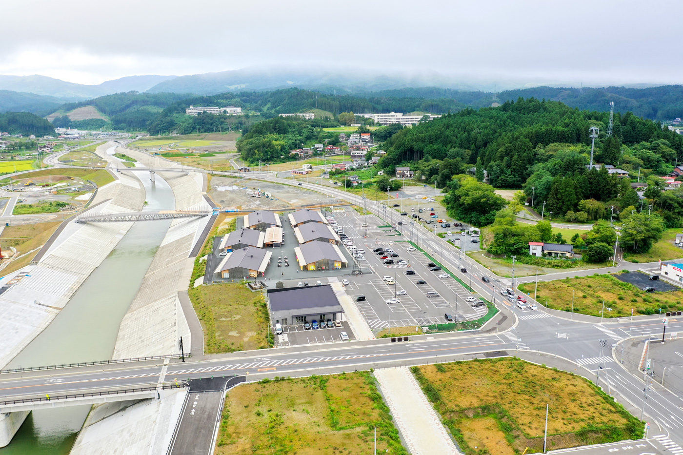 広々とした駐車場と複数の建物が並ぶエリアのすぐ隣を川が静かに流れており、その周囲には緑に覆われた山々と住宅地が広がっている自然と生活が共存する風景の写真