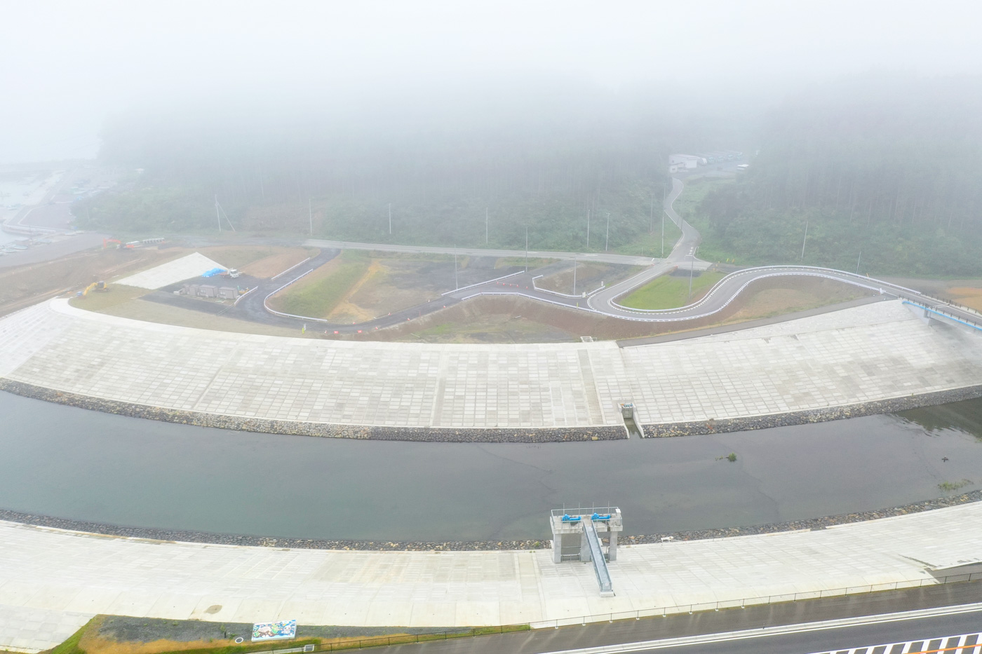 霧が立ち込める風景の中で、川沿いに護岸工事が施された区域が広がり、その背後には曲がりくねった道路と緑地帯が続いている様子を写した川沿いの風景の写真