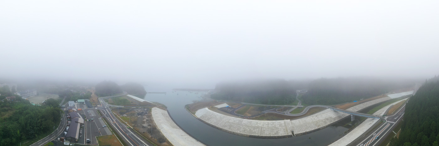 霧が立ち込める中、川が静かに流れており、その周囲には舗装された道路と複数の建物がぼんやりと見える幻想的な雰囲気の川沿いの風景の写真