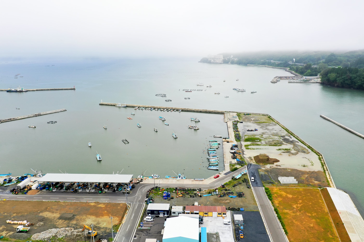 港の防波堤に囲まれた穏やかな海に多数の漁船が停泊しており、手前には倉庫や事務所のような建物が並ぶ港湾施設が広がっている漁港の写真