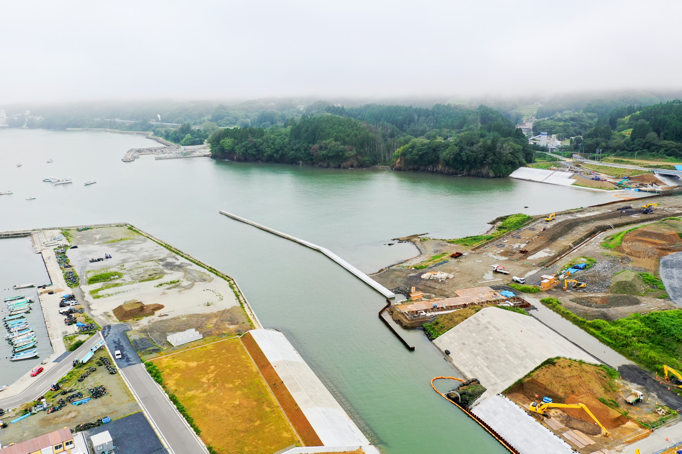 川がゆるやかに流れる風景の中で、その周辺に重機や資材が並ぶ工事現場が広がっており、整備中の道路や構造物が見える川沿いの工事現場の写真
