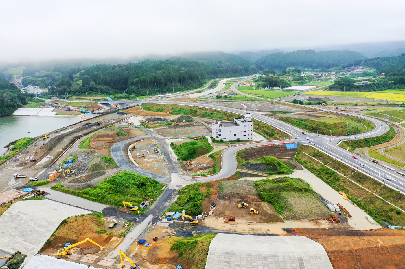緑に覆われた山々に囲まれた広大な建設現場で、複数の重機が稼働しており、整地された地面の中央には白い建物が建っている様子が見える建設現場の写真