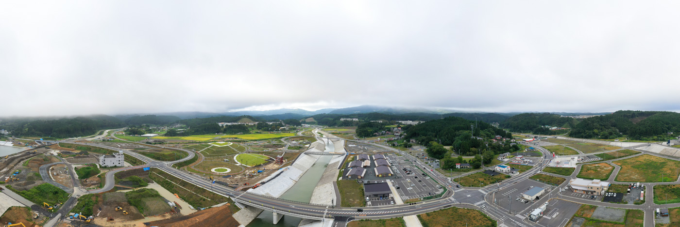 広々とした範囲にわたって道路が網の目のように走り、周囲には建物や緑地が点在しており、遠くの地平線には連なる山々が見える都市と自然が調和した風景の写真