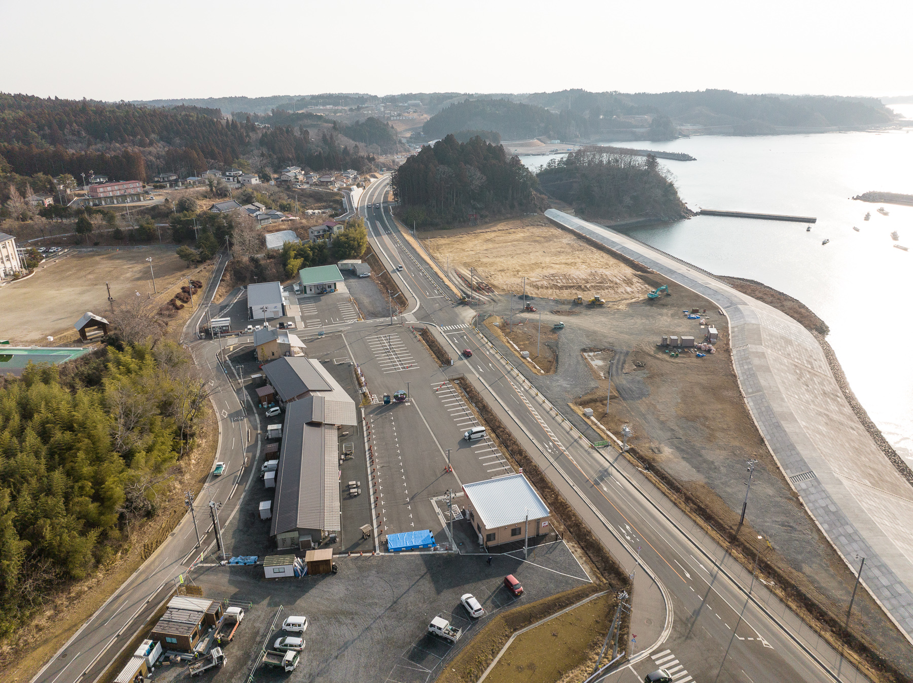 手前に複数の建物と駐車場がある道路沿いの施設が見え、その奥には海に向かって伸びる堤防と草地が広がっている昼間の風景を高台から定点カメラで撮影した写真