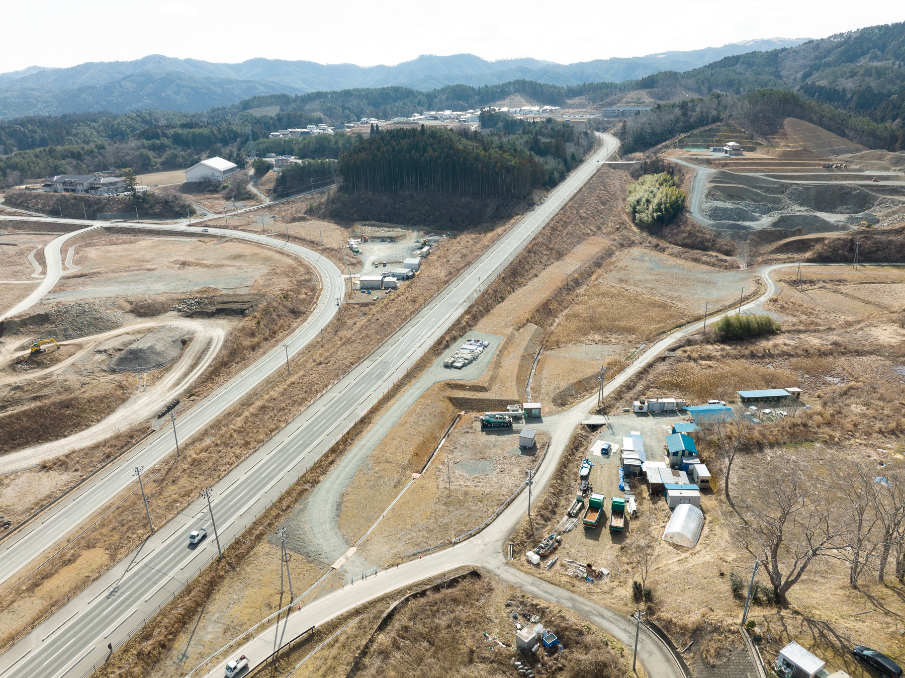 山に続く道路の両側に小さな施設や車両が並び、手前には農業用ビニールハウスのような建物も見える、内陸側の復興工事拠点付近の昼間の風景を高台から定点カメラで撮影した写真