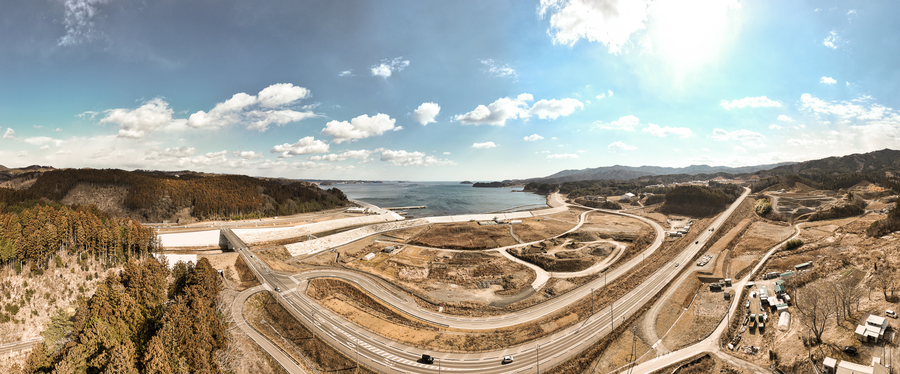 中央に大きく曲がりくねった道路が広がり、その先に広大な海と港、周囲に山々と森林が続く復興工事中の海岸地域の昼間の風景を高台から定点カメラで撮影した写真