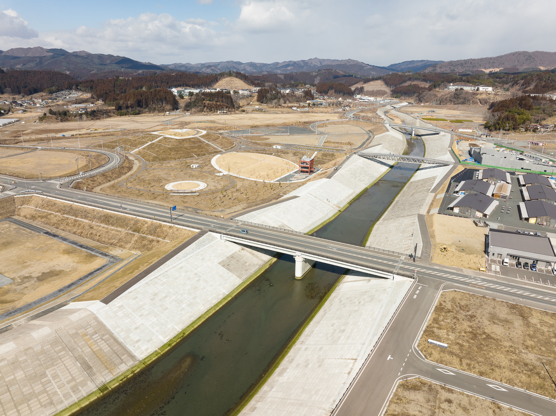 中央を流れる川に白い護岸が整備され、その両側に道路と造成地が広がり、少し離れた場所に新しい建物が並んでいる復興地域の昼間の風景を高台から定点カメラで撮影した写真