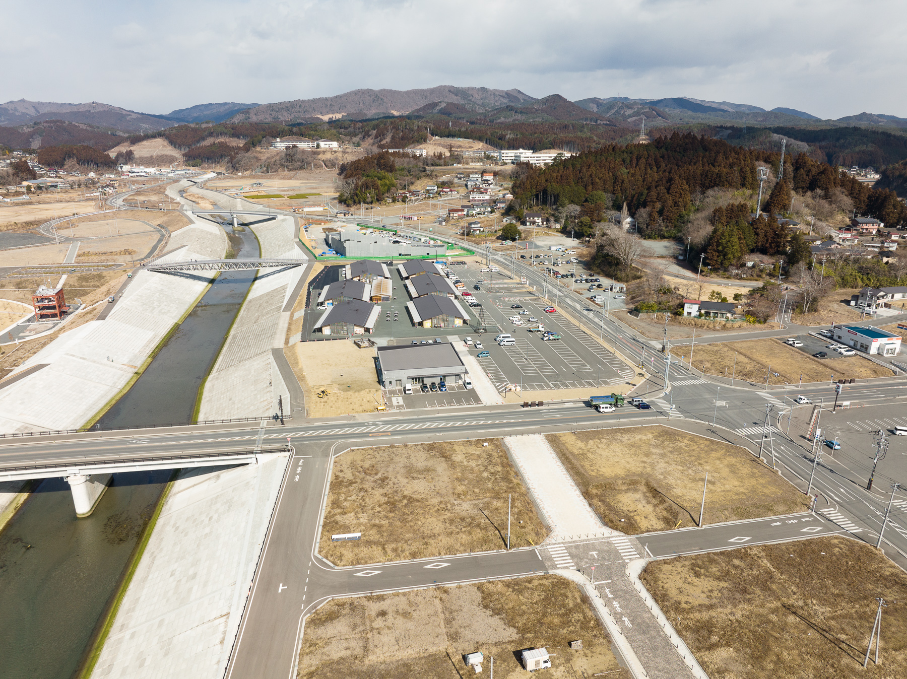 整備された堤防の右側に広い駐車場と商業施設があり、左奥には川と山が広がる街の再開発エリアの昼間の風景を高台から定点カメラで撮影した写真