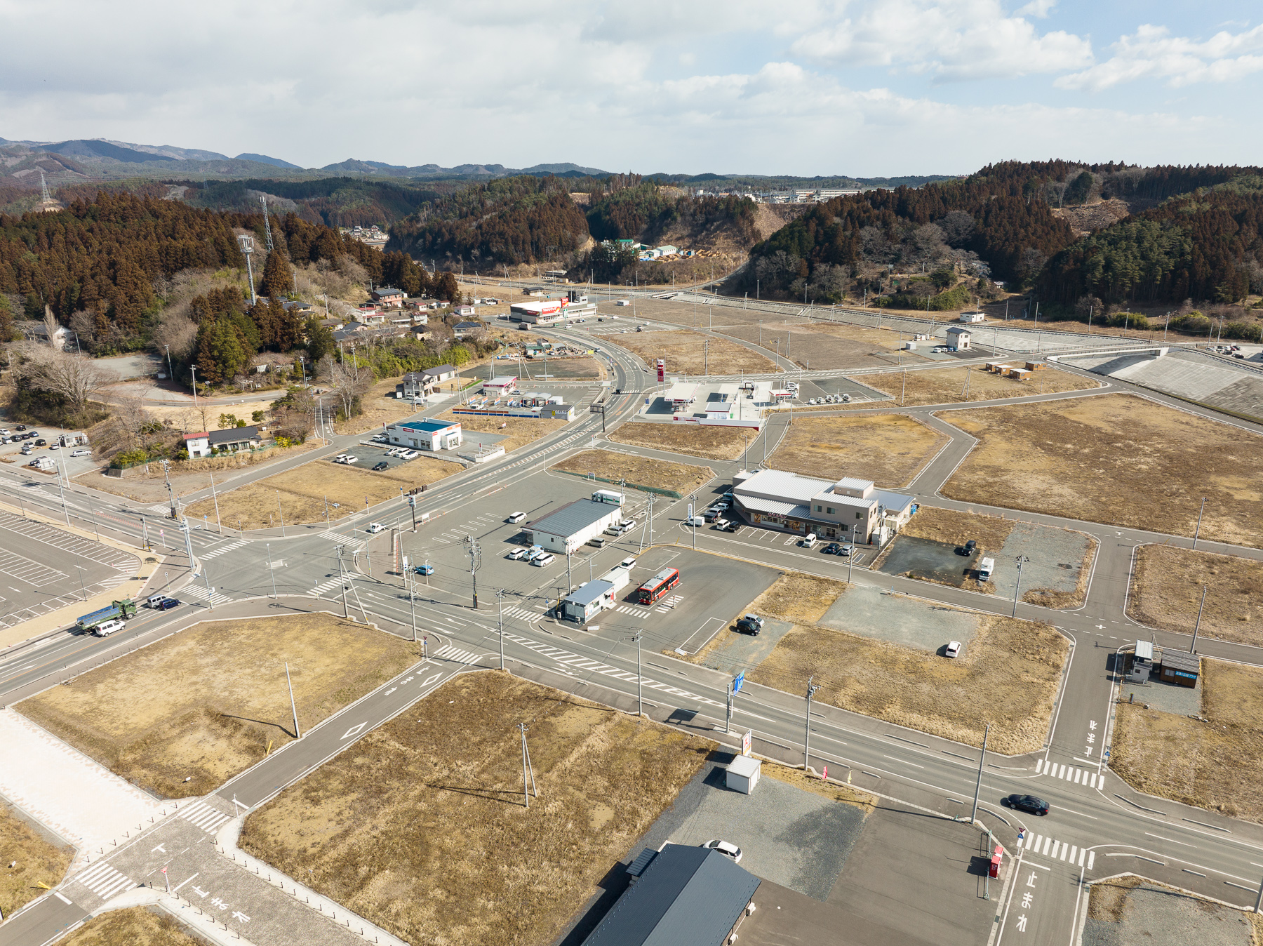 交差点に面してコンビニや事務所があり、その周囲に空き地と新しいインフラが整備された小さな町の中心部の昼間の風景を高台から定点カメラで撮影した写真