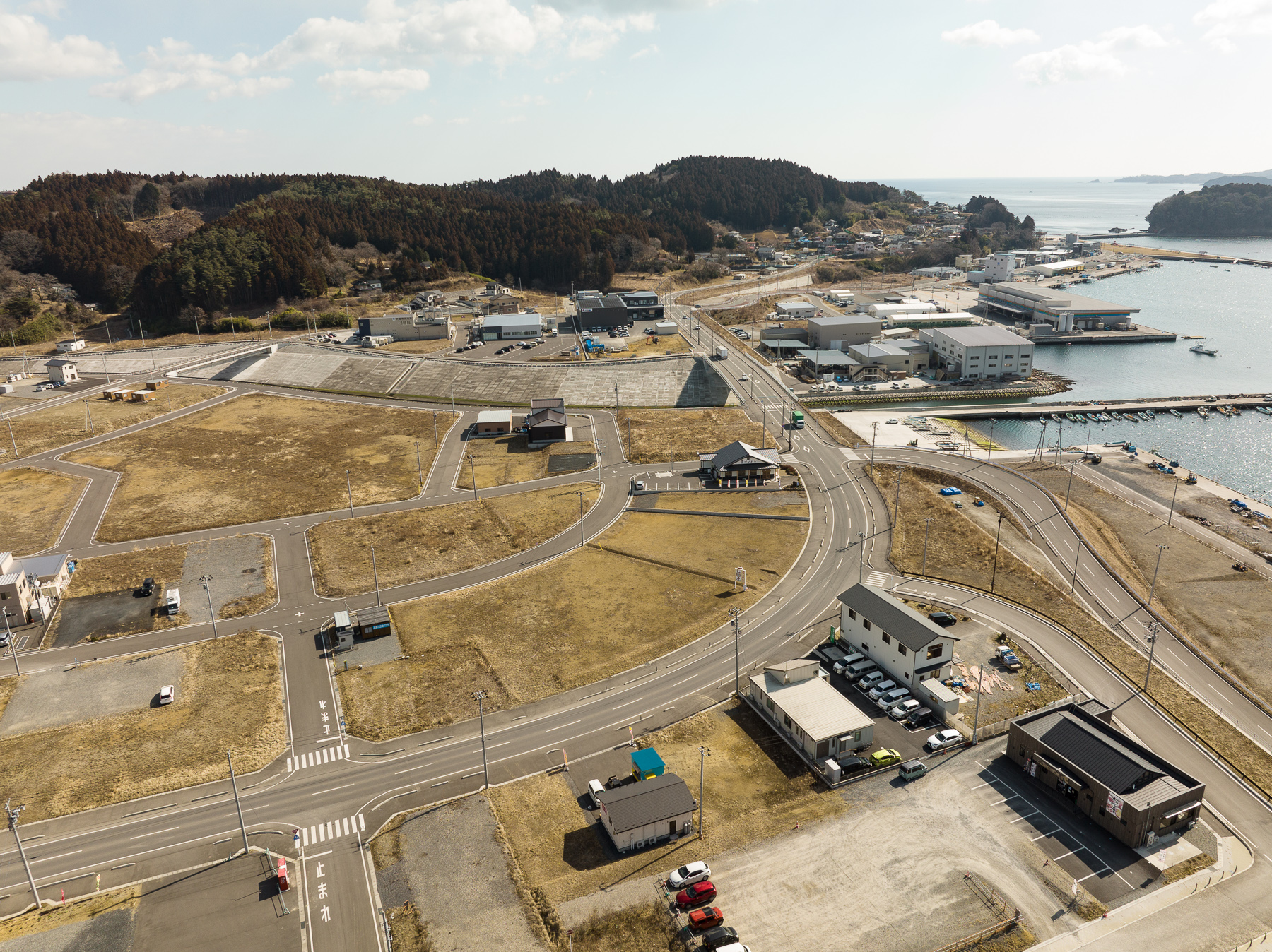 緩やかなカーブを描く道路に囲まれた空き地が広がり、その先に港湾施設や倉庫が並ぶ沿岸部の再開発エリアの昼間の風景を高台から定点カメラで撮影した写真