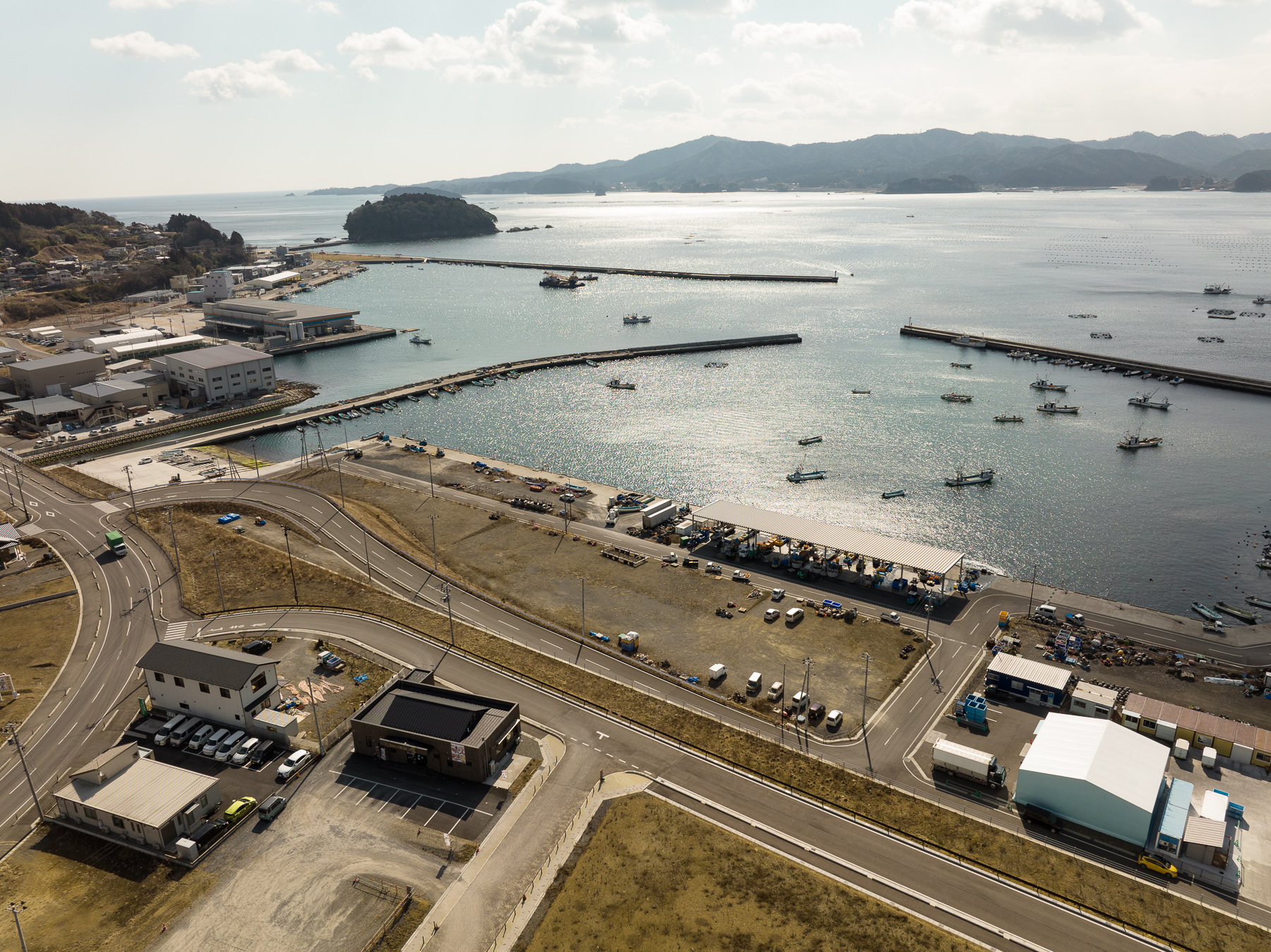 複数の漁港施設と倉庫が湾に面して立ち並び、奥に小島と広がる海が見える漁港地域の再整備後の昼間の風景を高台から定点カメラで撮影した写真