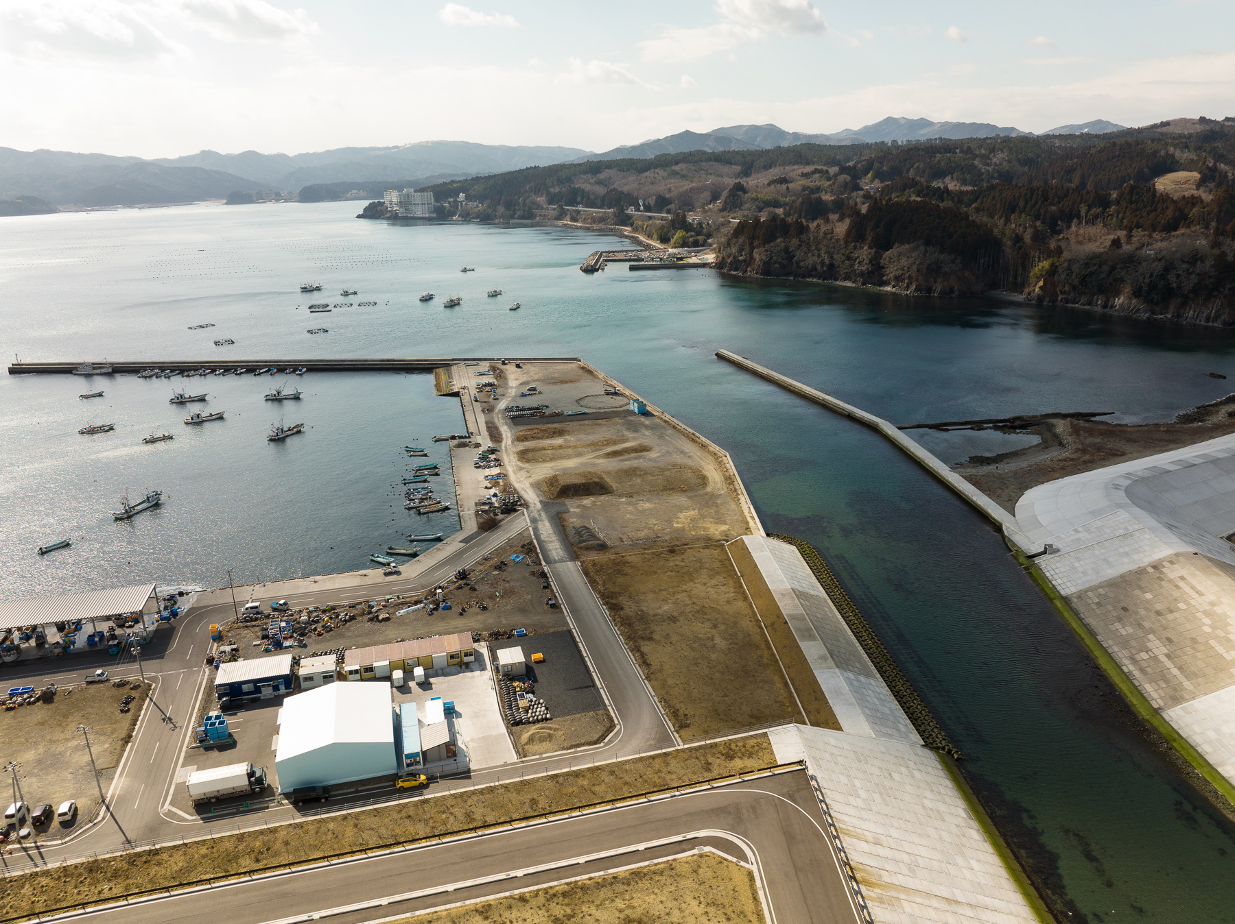 波の穏やかな湾に沿って船が並び、岸には作業場と空き地があり、右に整備された川の流出口が見える港町の昼間の風景を高台から定点カメラで撮影した写真