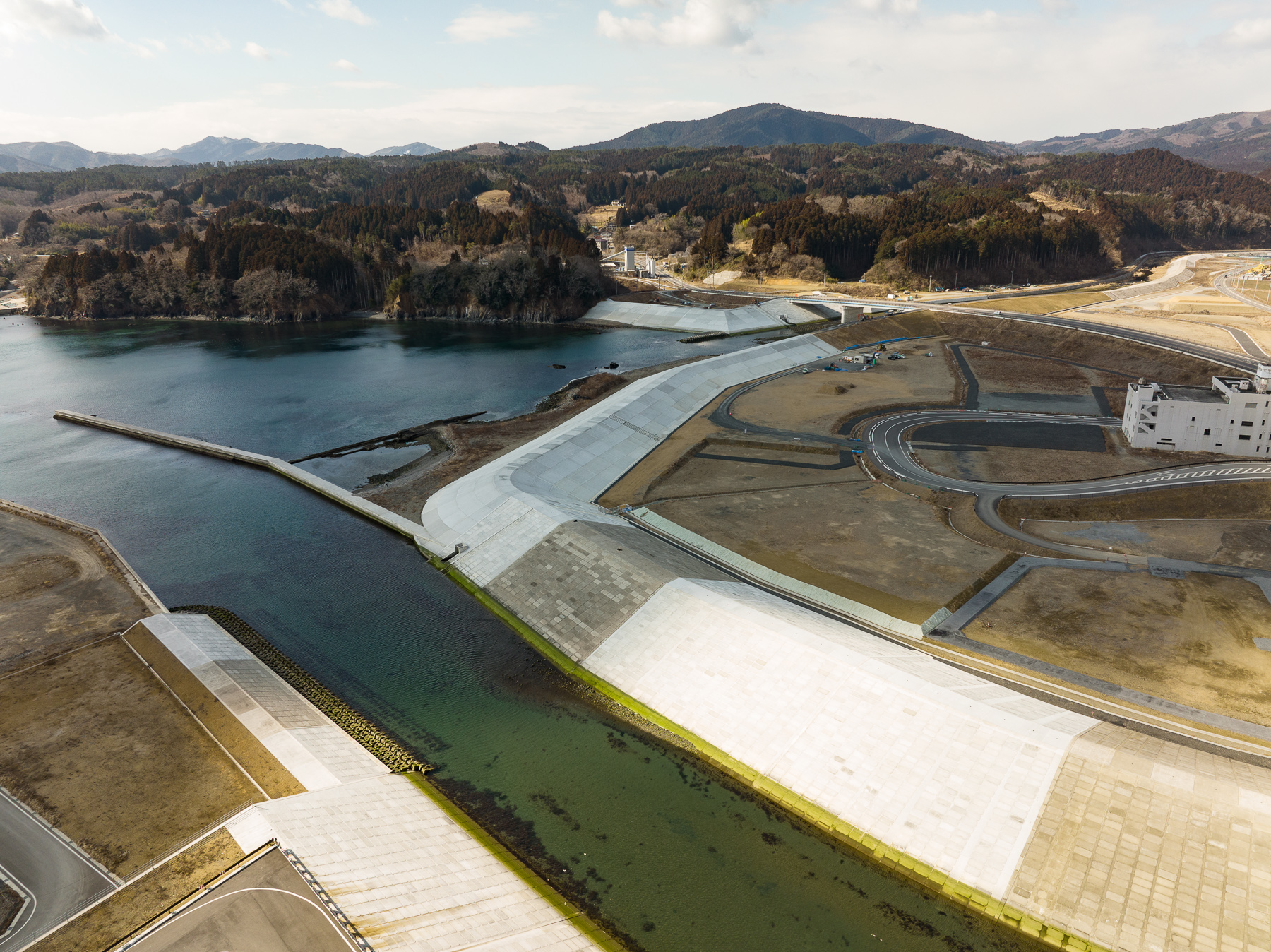 緑色がかった海の入り口にコンクリートの堤防が設けられ、右側にカーブを描く道路と建物がある港湾整備区域の昼間の風景を高台から定点カメラで撮影した写真