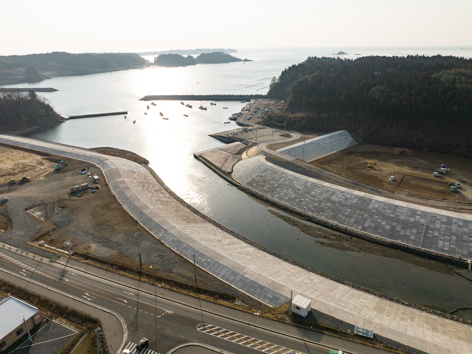 手前に湾曲した護岸が広がり、その先に海と漁船が浮かぶ港が見え、山に囲まれた入り江のような昼間の風景を高台から定点カメラで撮影した写真